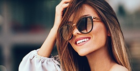 A young woman wearing sunglasses and smiling with greater confidence because of her veneers